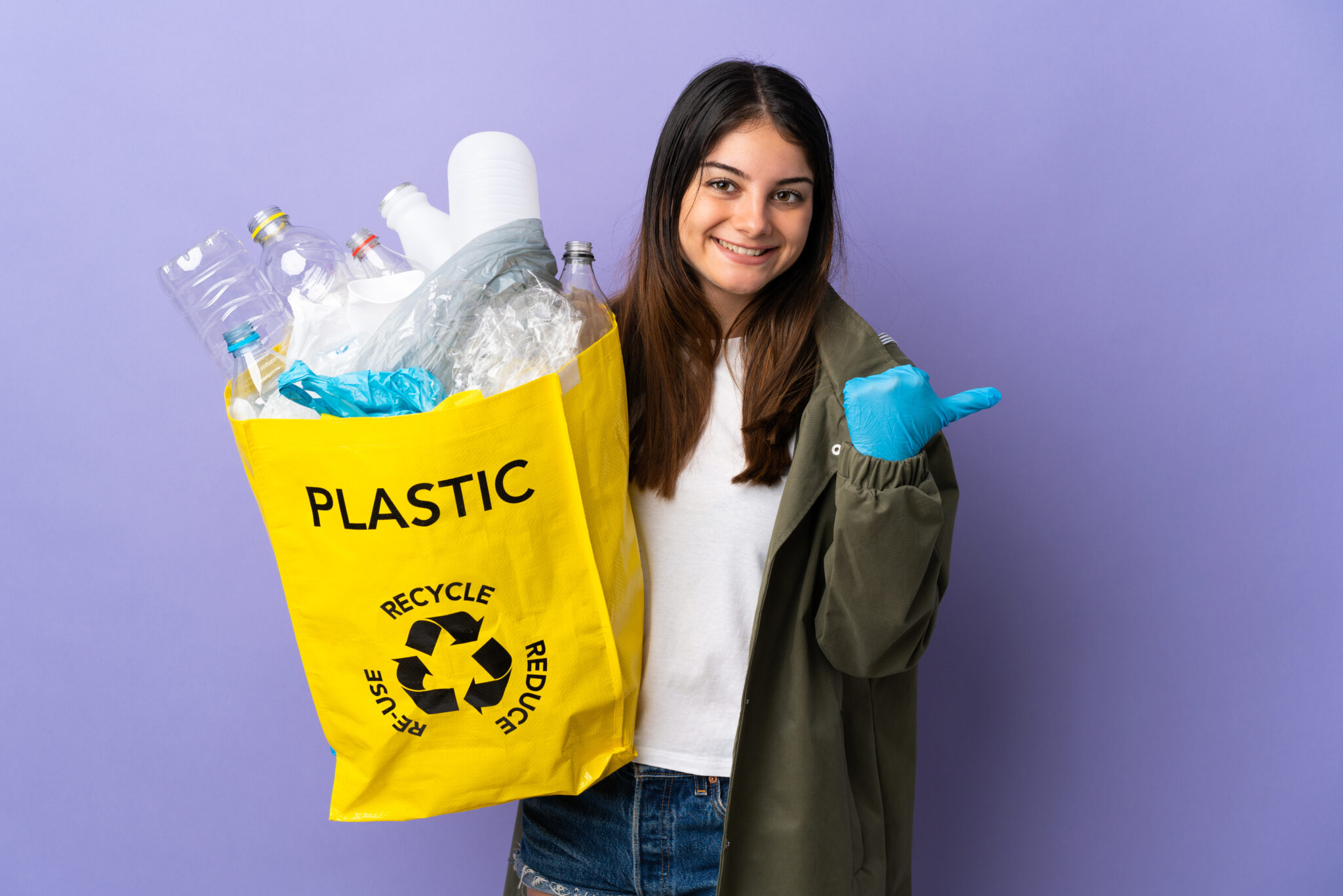 Young woman holding a bag full of plastic bottles to recycle isolated on purple background pointing to the side to present a product